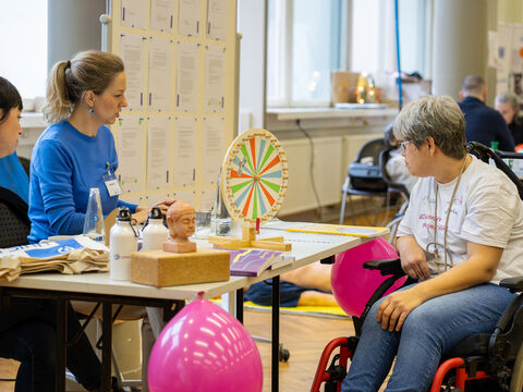 Zwei Mitarbeiterinnen der Lebenshilfe Bildung zeigen einer Rollstuhlfahrerin ein Glücksrad für ein Gewinnspiel. 